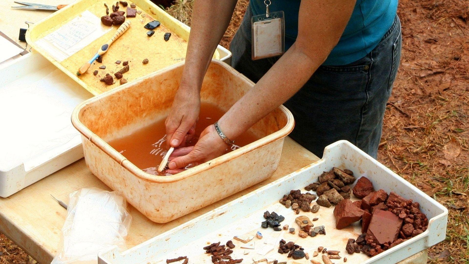 An archeologist cleans a tool while outside near other tools and dirt.