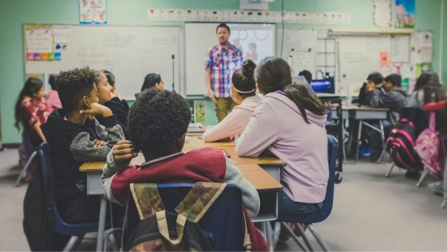 This photo shows a group of diverse children sitting at desks observing a teacher who is standing at the front of a classroom
