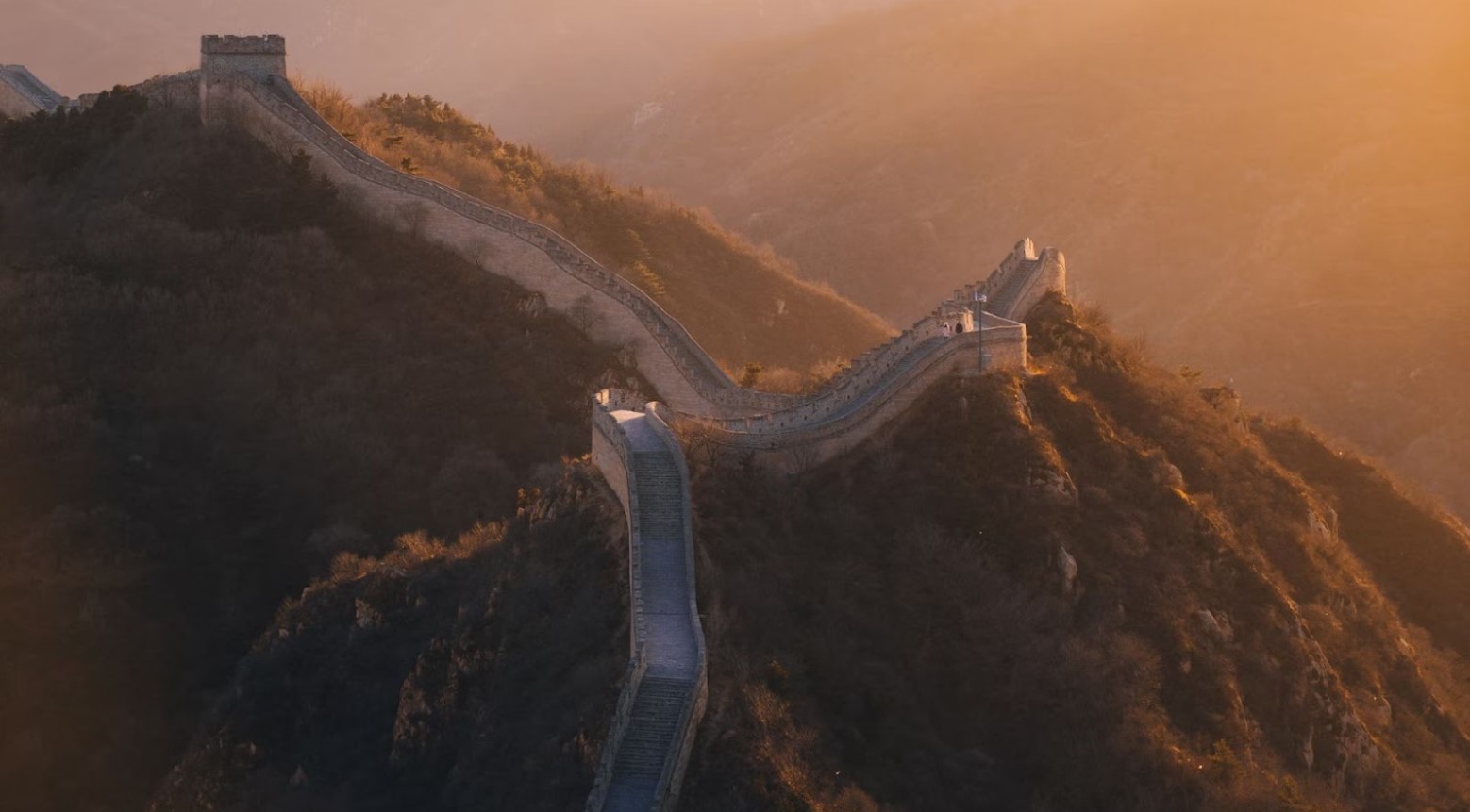The Great Wall of China at sunset. The wall goes up a hill and then curves around to the left.