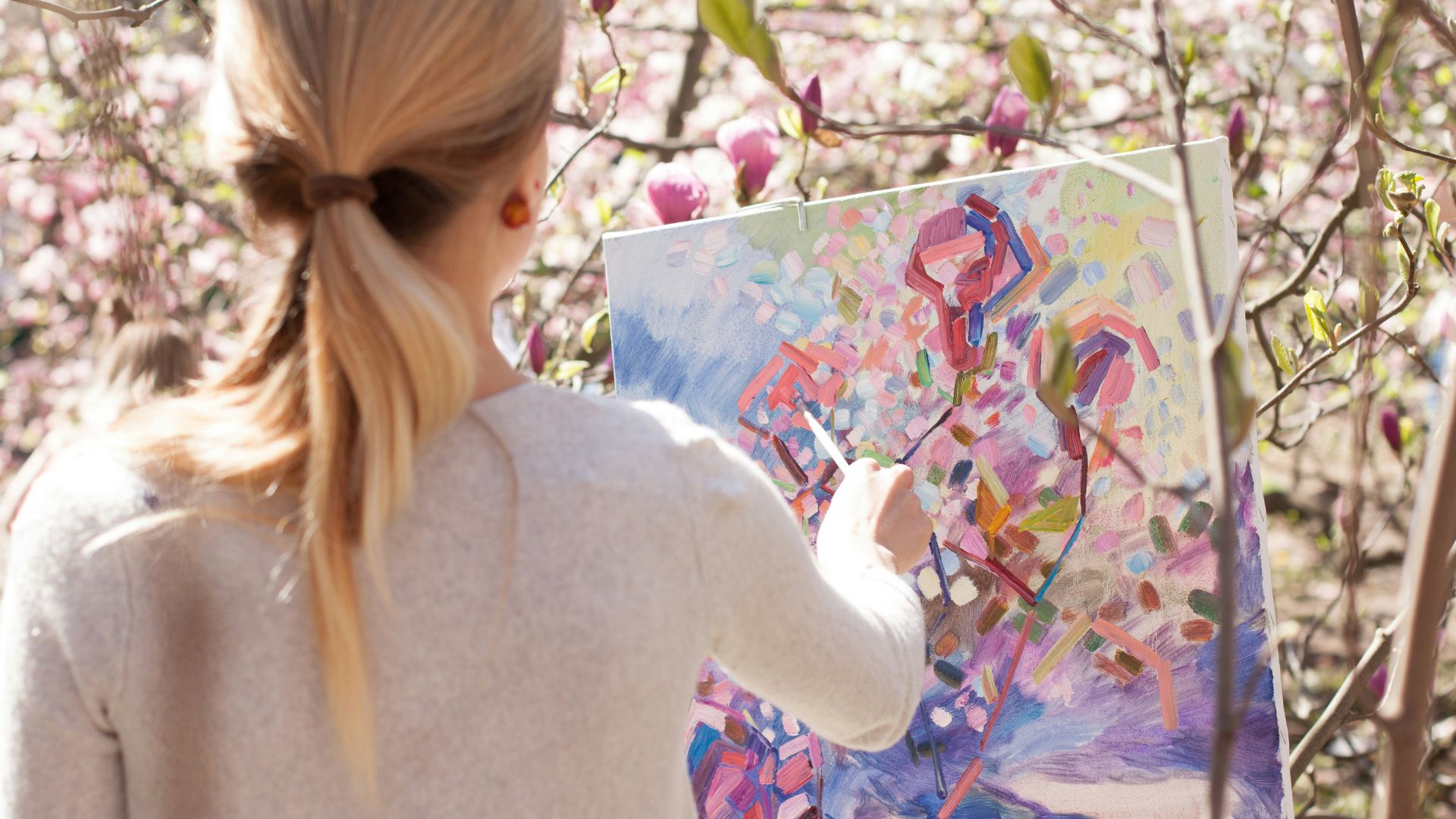 A blonde woman stands in front of a canvas and paints on it while outside.