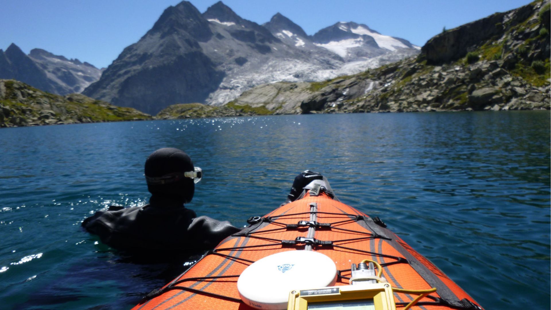 View from a kayak on a clear mountain lake, with the front of the bright orange kayak stretching out into the frame. A person dressed in black with a snorkeling mask on their head is sitting at the rear, gazing towards the sharp, snowy mountain peaks in the distance