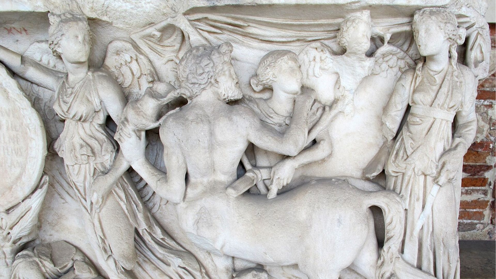 Close-up of an ancient Roman marble relief sculpture. The scene depicts figures in high relief, including a central male figure whose back is turned to the viewer, and several surrounding characters who appear to be in a dynamic interaction with him and each other