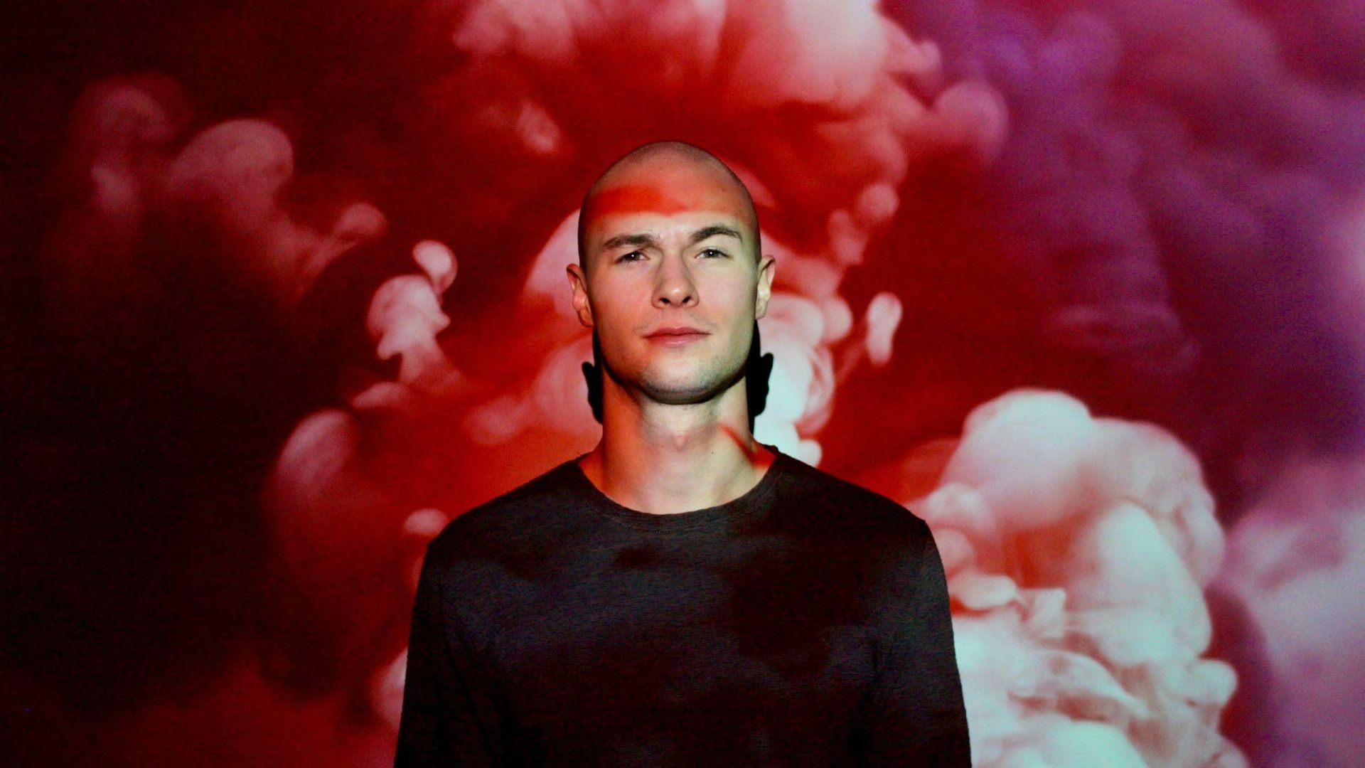 A bald man standing in front of a red and pink background.