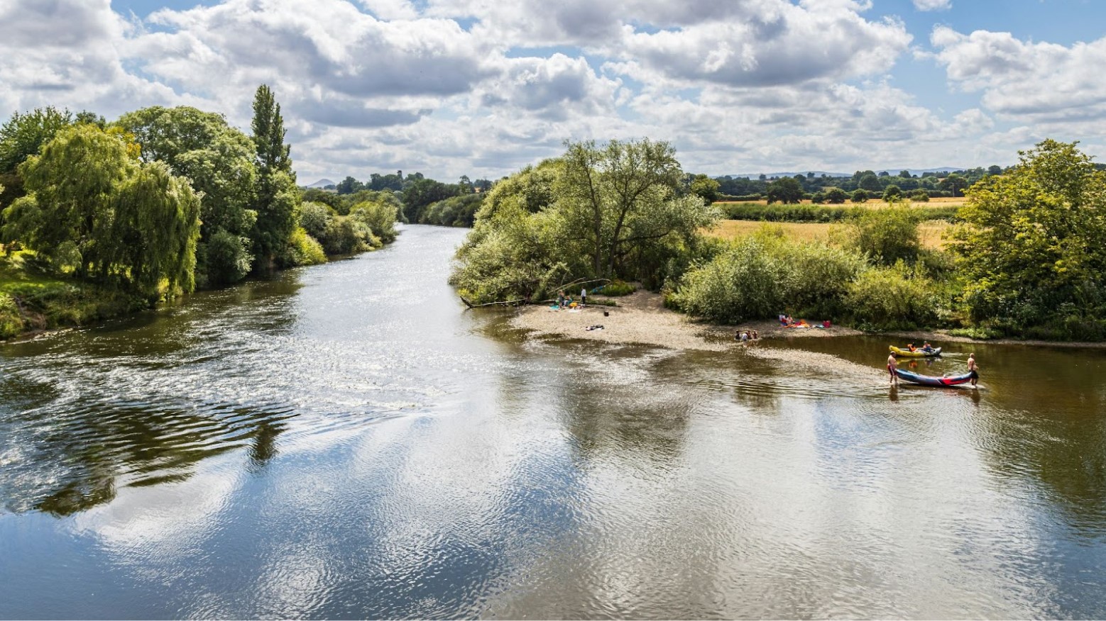 A group of people in the water by a river in England, with green trees and fields in the distance.