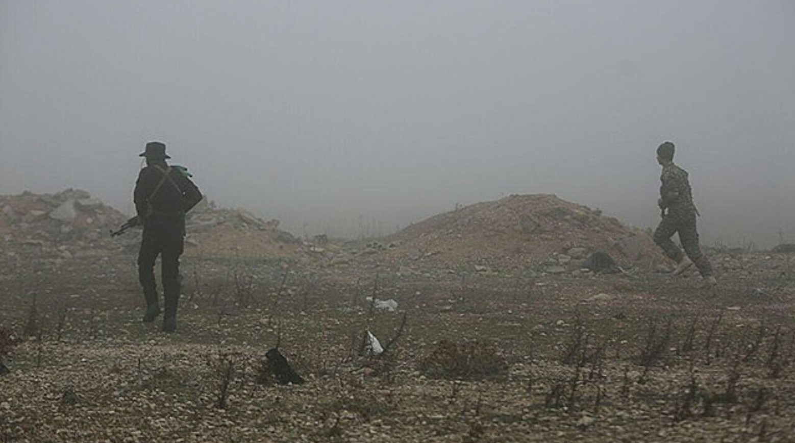 An image from the Syrian War. Two soldiers are walking and the land they are walking on is mostly rubble. Some parts are flat and others are mounds of rubble.