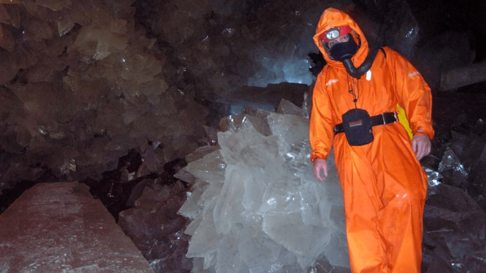 An explorer in an orange suit with an oxygen tank poses next to a massive gypsum crystal.