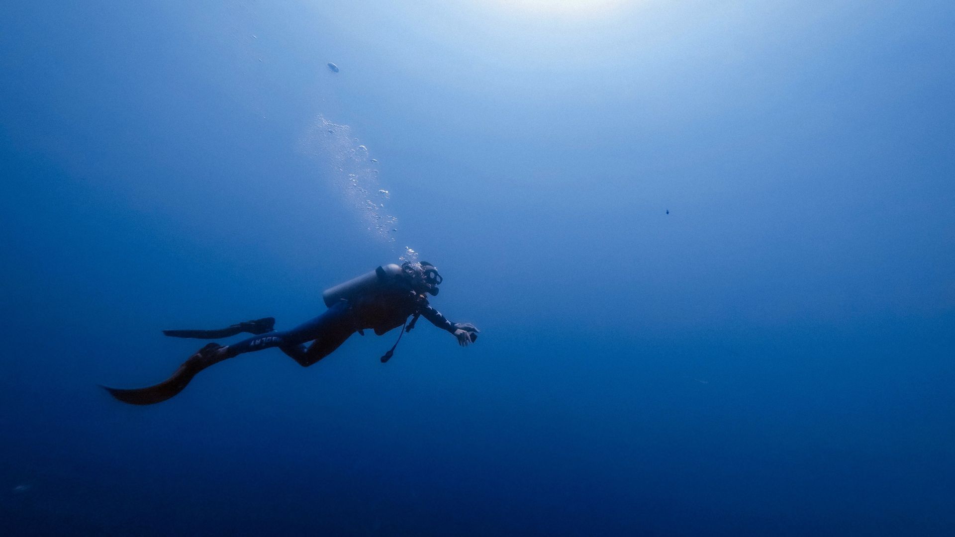 A lone scuba diver is immersed in the vast blue expanse of the ocean. The diver, equipped with a large tank, fins, and a dive mask, is oriented diagonally in the frame