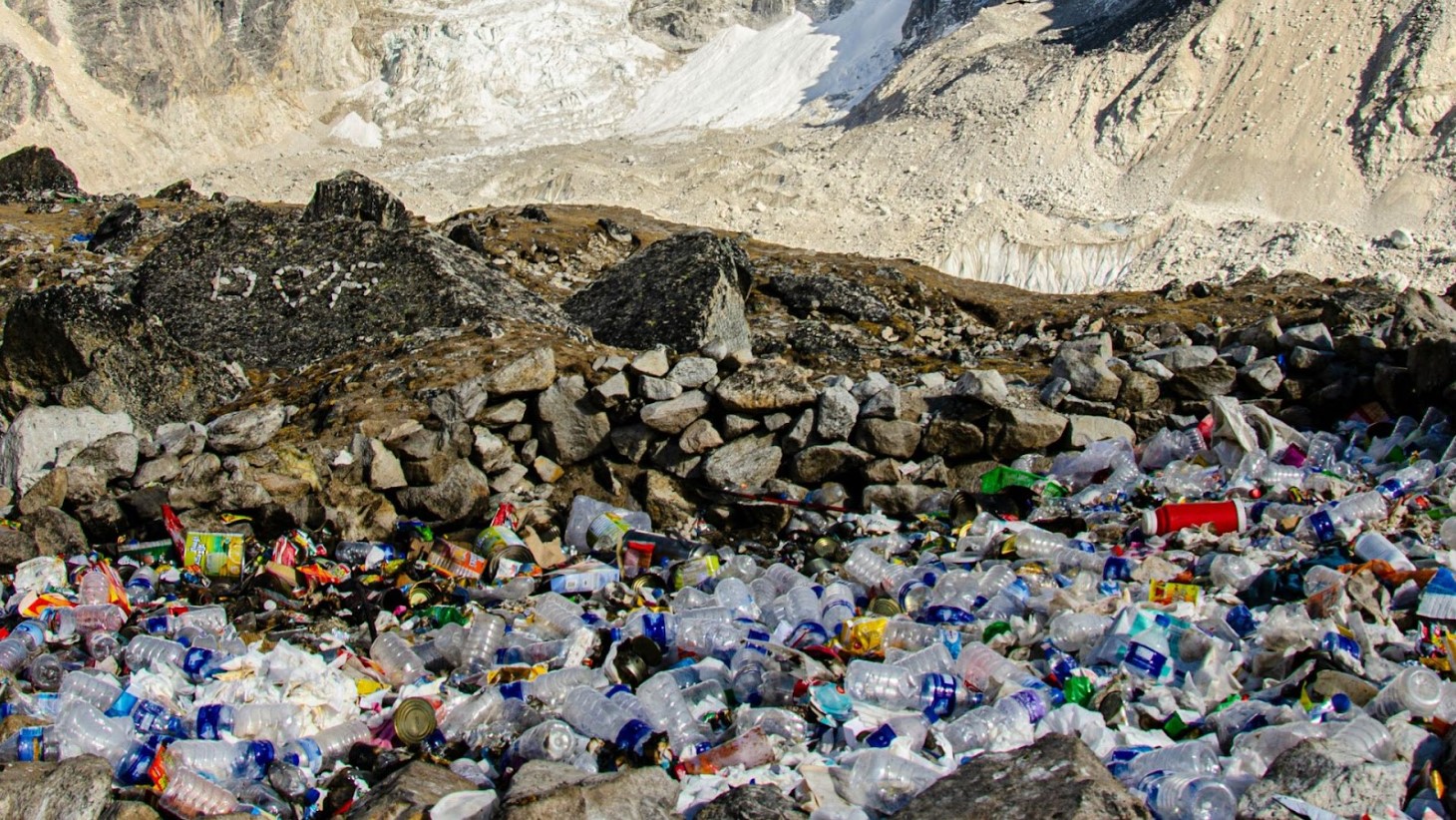 A stark contrast between nature and pollution is depicted in this image, where a large accumulation of colorful plastic waste covers the ground in a mountainous area. The waste, comprising of various bottles, wrappers, and other discarded items, is set against a backdrop of rocky terrain