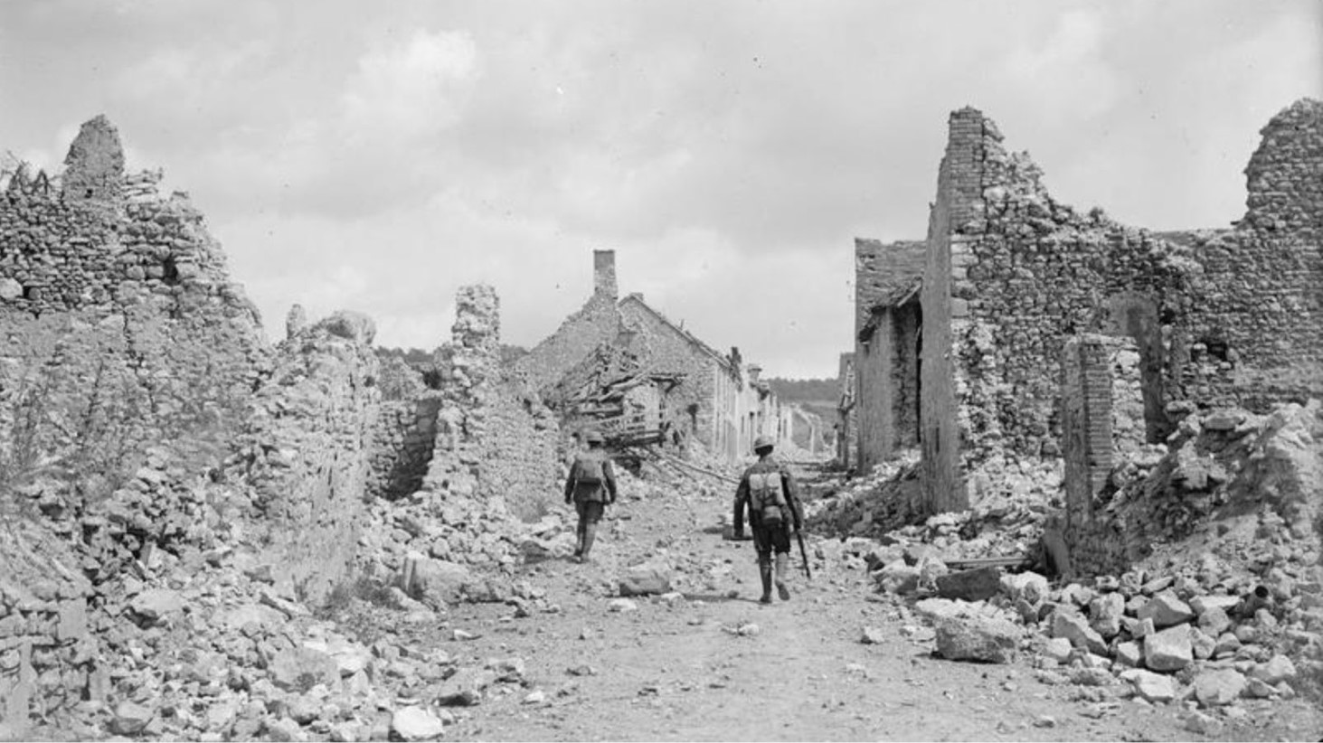 Black and white photograph of two soldiers walking down a desolate street lined with the crumbling ruins of buildings destroyed in wartime