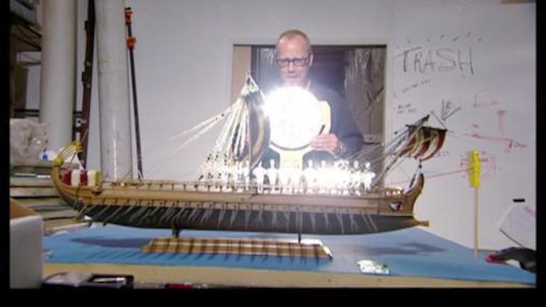 A man holding a bright light up to a model of an ancient Roman ship