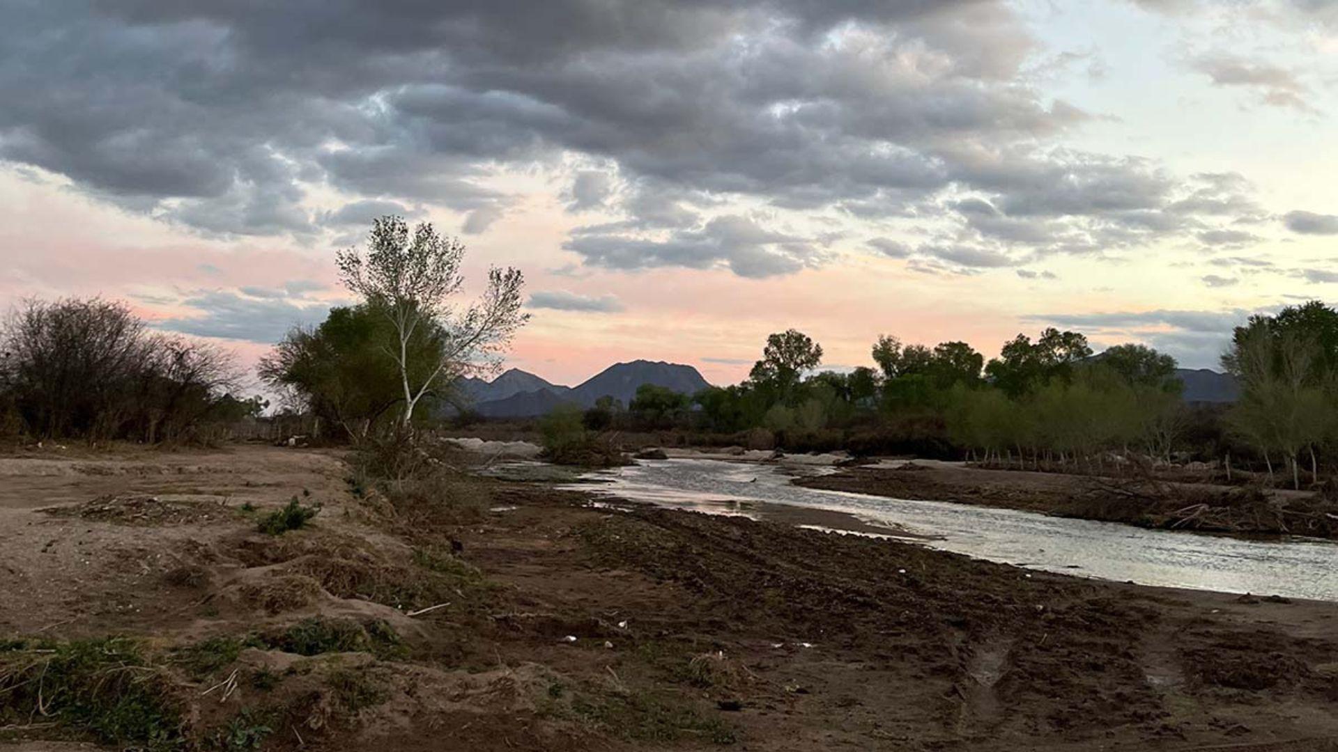 The image shows a scene of a desert riverbed at dusk, with gentle water flowing through. The foreground shows sandy banks leading to sparse vegetation and trees, while the background features silhouetted mountains