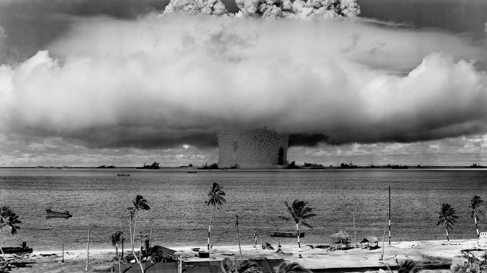 A grayscale photo of a towering mushroom cloud explosion off the coast of a beach.