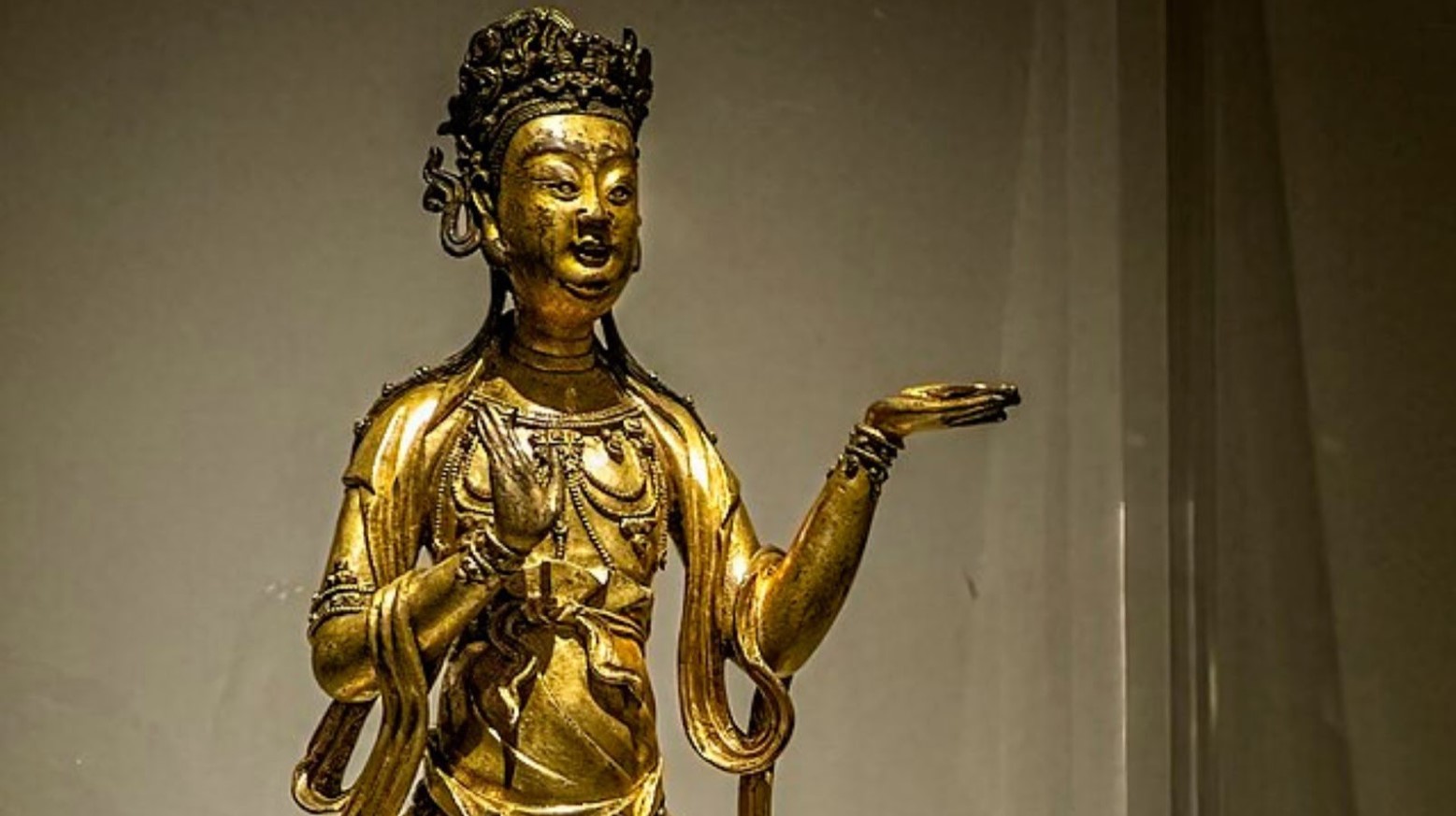A golden statue from the Ming Dynasty. The status is holding one hand to the side while the other is against the body. It has a crown on its head.