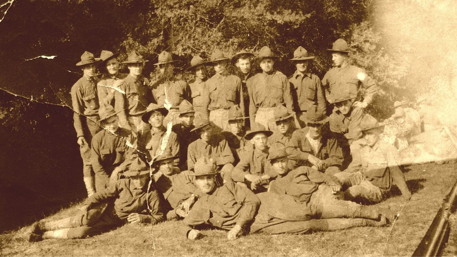 Sepia-toned photograph of a group of World War I soldiers. They are casually posed in two rows, some standing and others sitting or reclining on the grass. Each soldier is wearing a uniform and a brimmed helmet