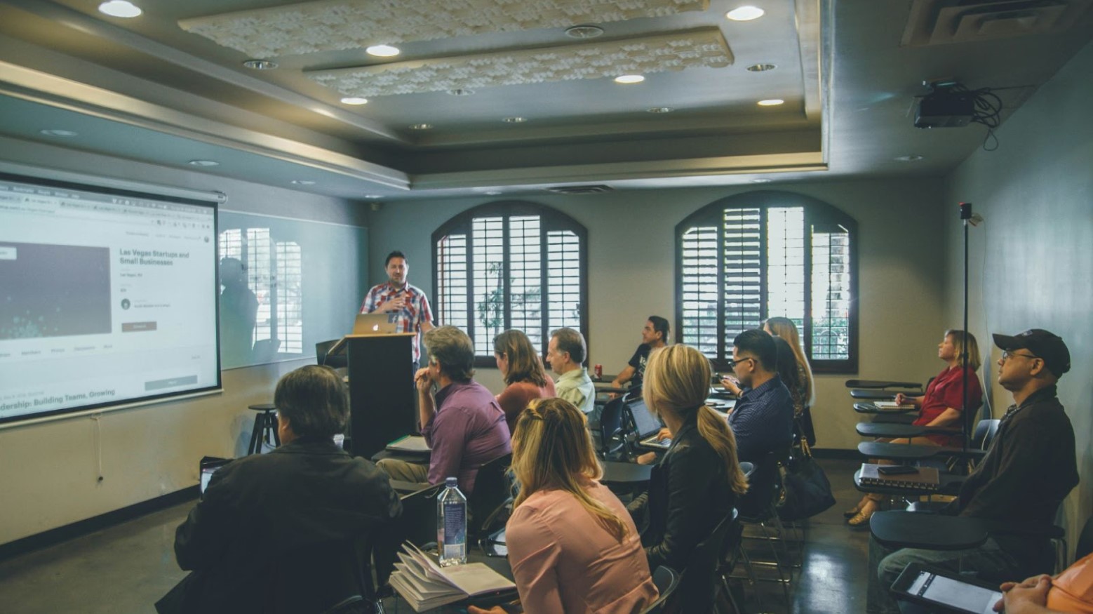 A classroom with a man at the podium and a powerpoint presentation is shown. Many students of all ages direct their attention towards the screen