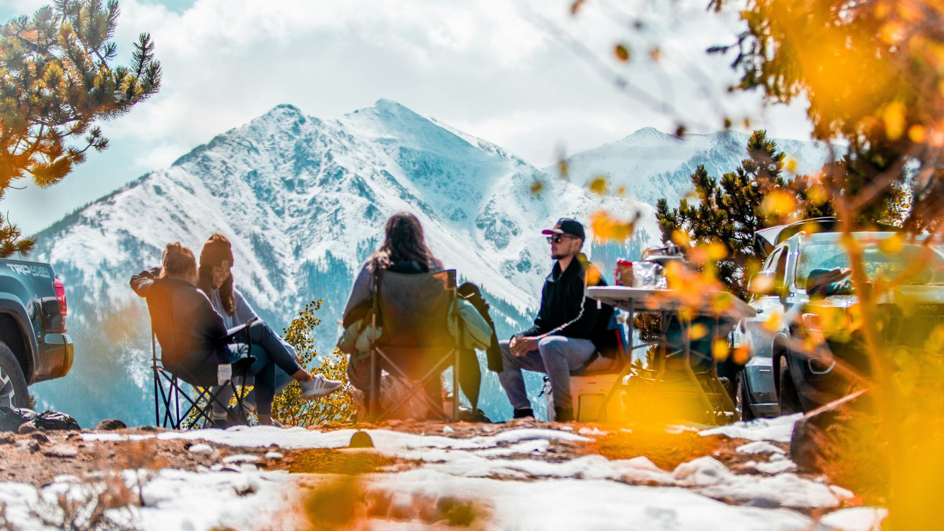 Friends sitting outside in chairs while camping in front of a snowy mountain.