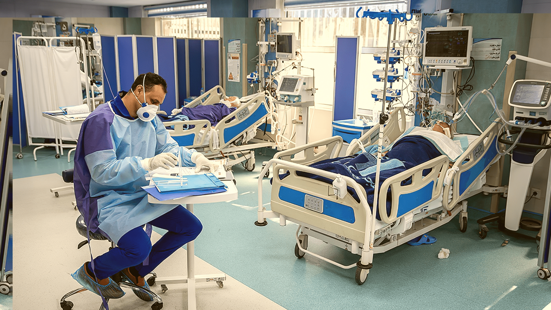 A doctor sits in a room with two patients monitored in beds.