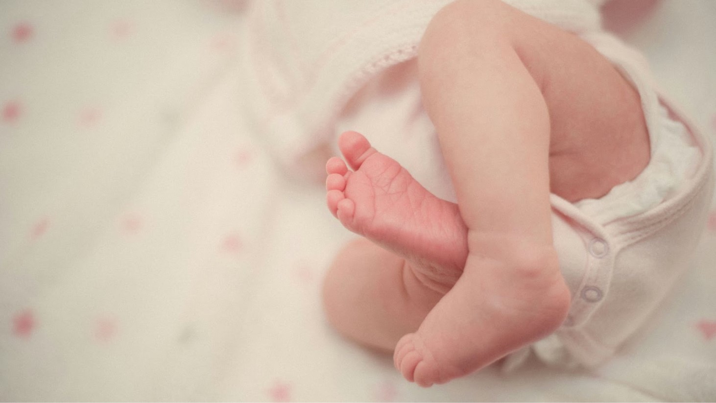 Close-up of a newborn baby's tiny feet with toes, wearing a white onesie, against a soft pink background with white hearts