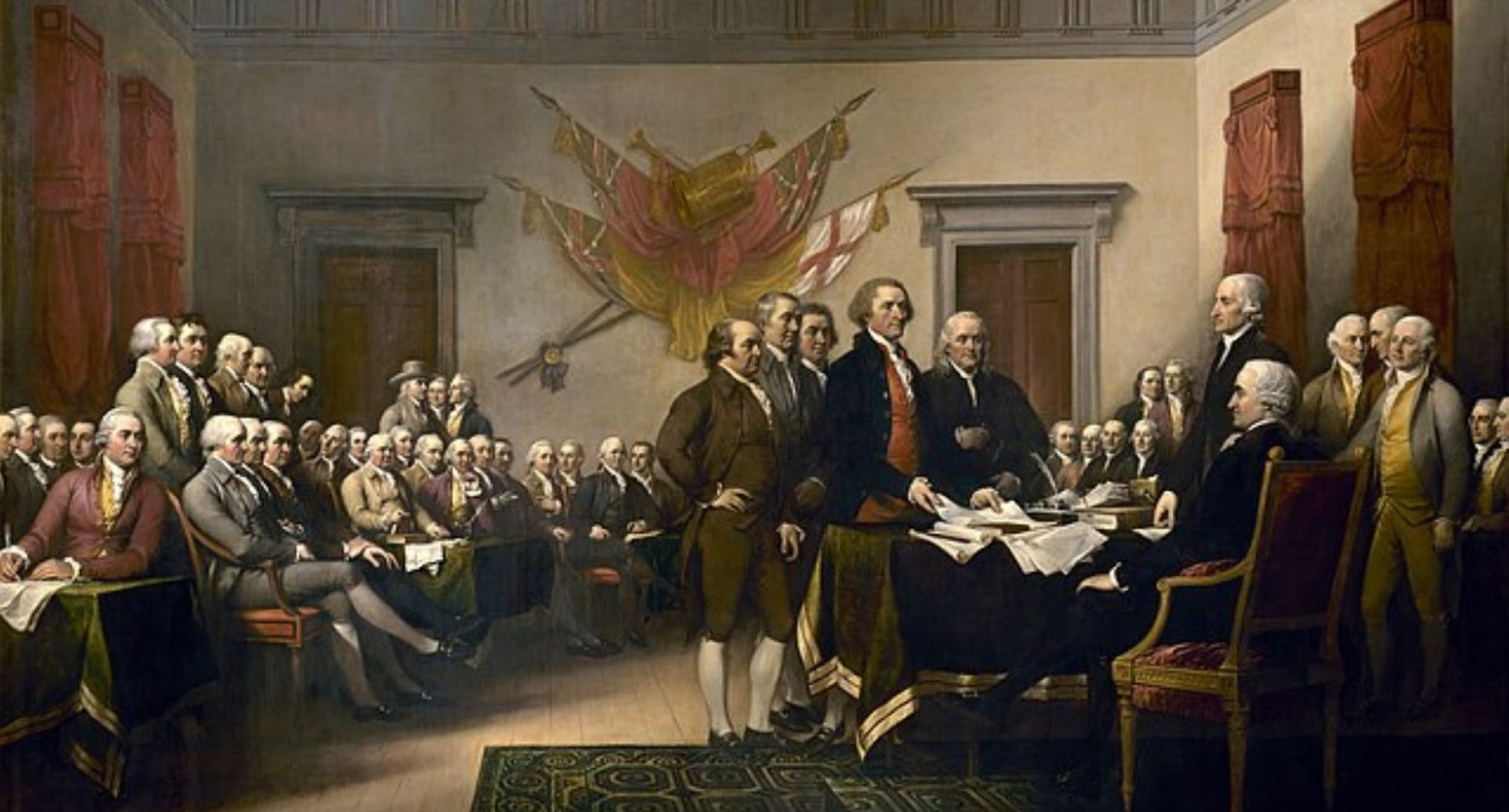 A scene depicting the Declaration of Independence. There are five men approaching a desk with a man sitting behind. There is a crowd of people also in the room who are sitting.