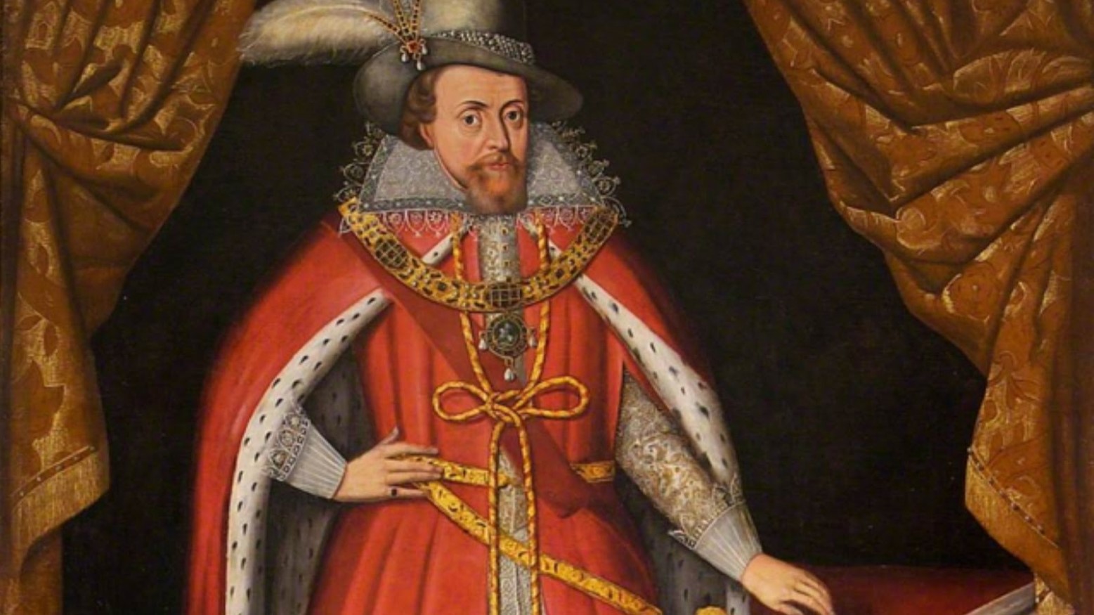 King James I wearing royal state clothes. He is wearing a red tunic and cloak that has a gold chain around it. He is also wearing a grey hat with a feather coming out of it. He is standing between two open golden curtains.