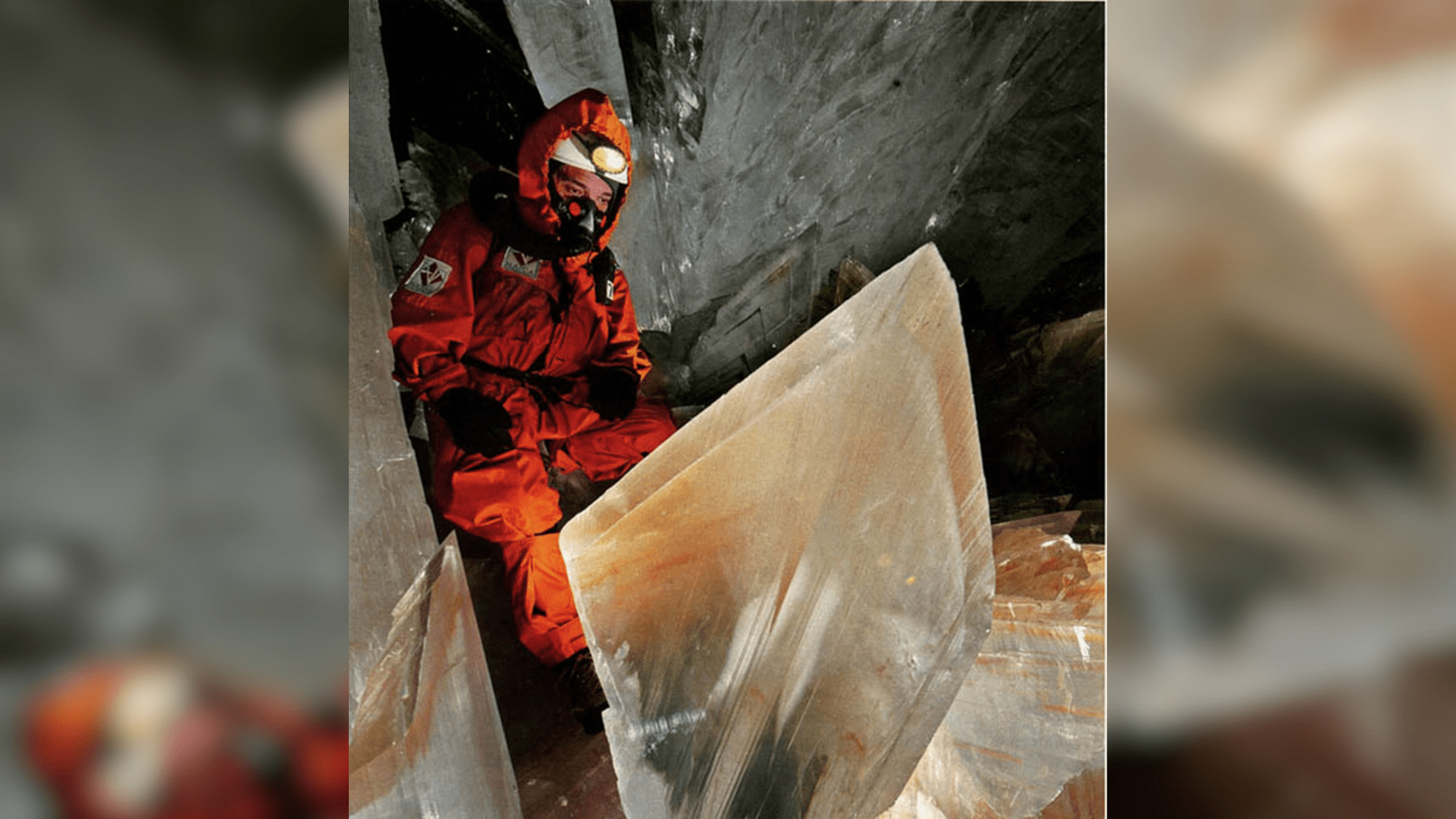 An explorer in an orange safety suit and an oxygen mask sits in the Cave of Swords looking at a giant gypsum crystal.