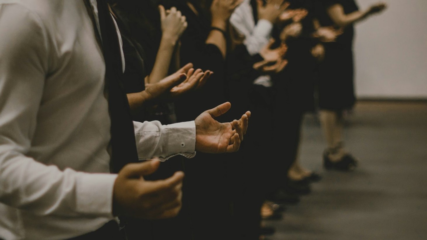 A side view of a row of people dressed in formal attire, with their hands palm up. The photo captures a range of individuals in a moment of worship