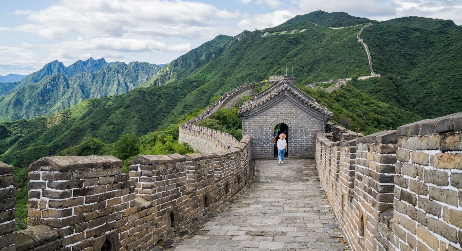 The Great Wall of China. The photo is taken from inside the wall and shows a person walking ahead. The wall can be seen going up a hill in the distance.