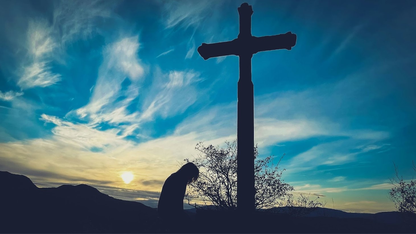 Silhouette of a person kneeling in prayer before a cross during sunrise or sunset. The cross and individual are outlined against a vibrant blue sky with wispy clouds