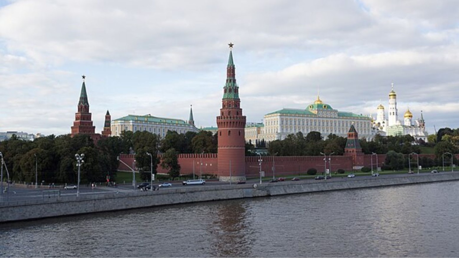 The Moscow Kremlin. The walls are red with turrets that have a green top. The building is white, with a green roof and gold on top. It is positioned next to the water.