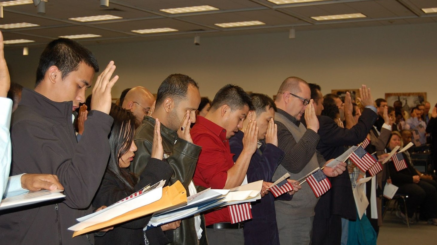 A group of individuals from diverse backgrounds standing and raising their right hands in a room during a naturalization ceremony