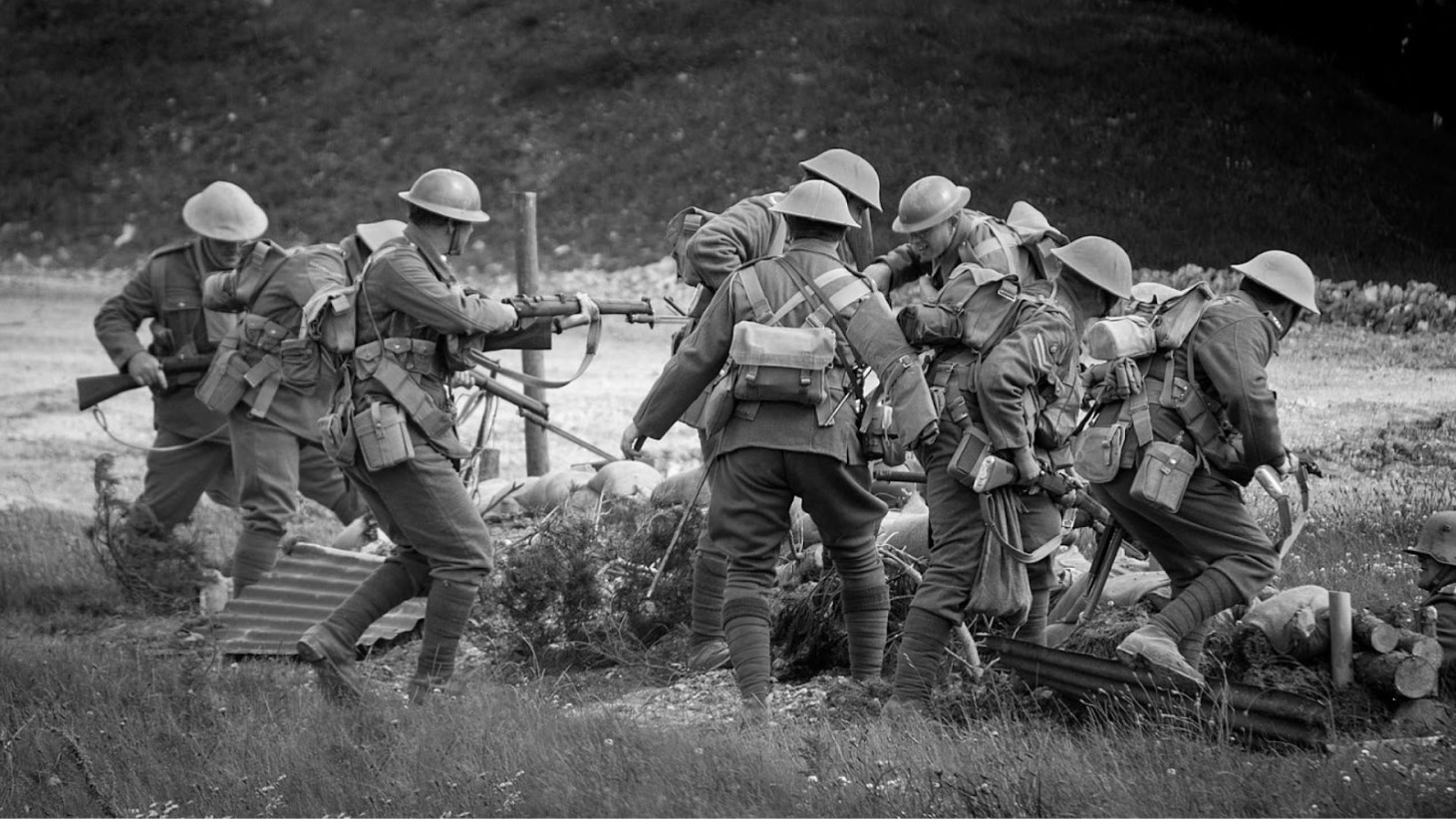 Black and white photograph of World War I soldiers. Several men in period military uniforms are positioned around a machine gun. They are wearing combat helmets and carrying backpacks, with one soldier operating the gun, others assisting with ammunition, and one lying prone as a lookout
