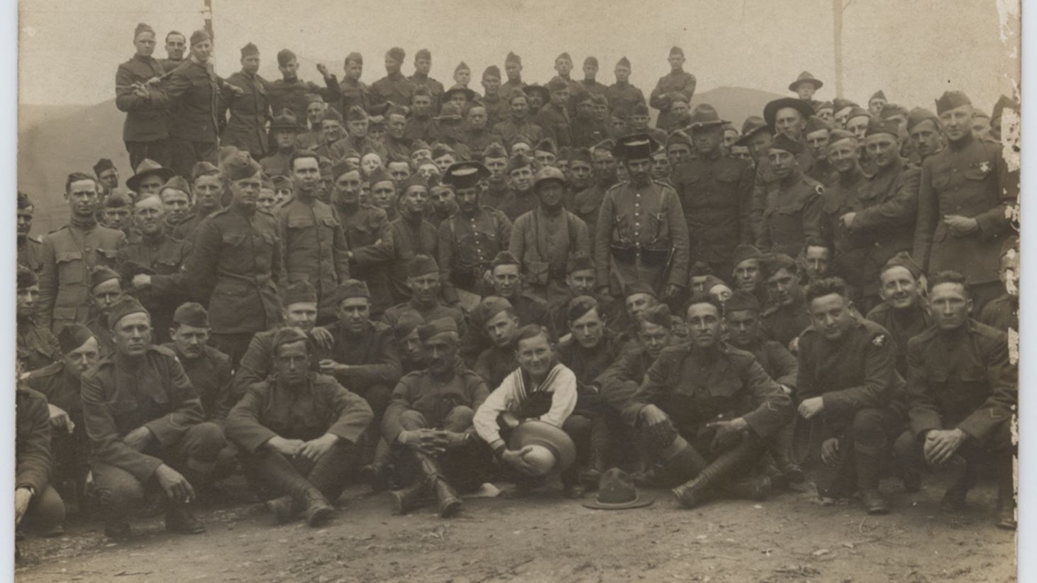 Sepia-toned historical photograph featuring a large group of World War I soldiers. They are gathered outdoors, with some seated in front, and others standing in multiple rows behind them