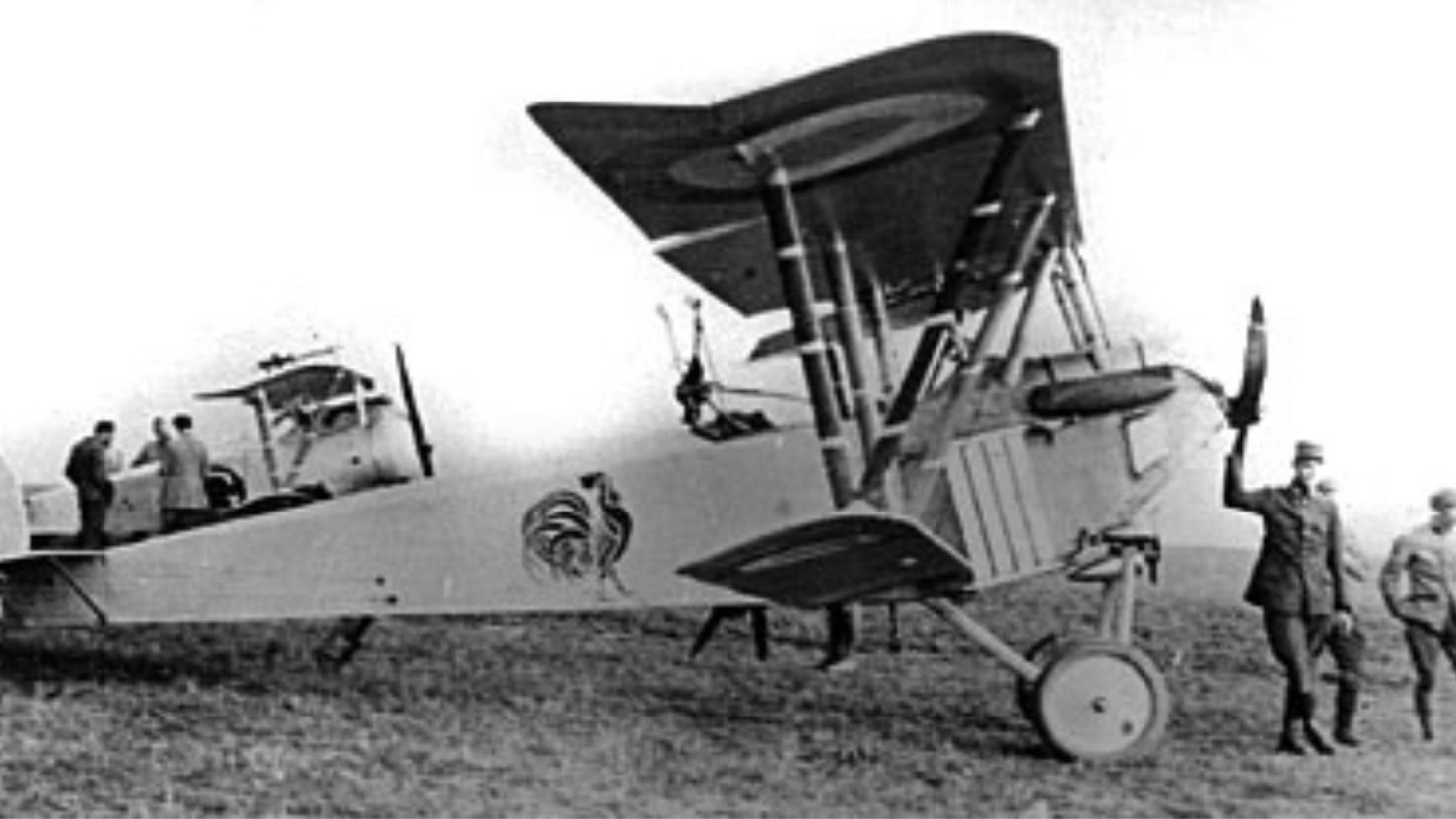 A black and white photo of an early biplane from World War I on a grassy field. The aircraft features a distinctive painted emblem on the side
