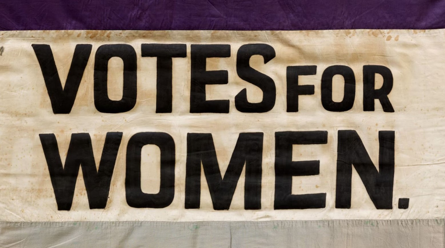 A Votes For Women flag. The flag is purple, white, and green; on the white part, it says “Votes For Women” in black writing.