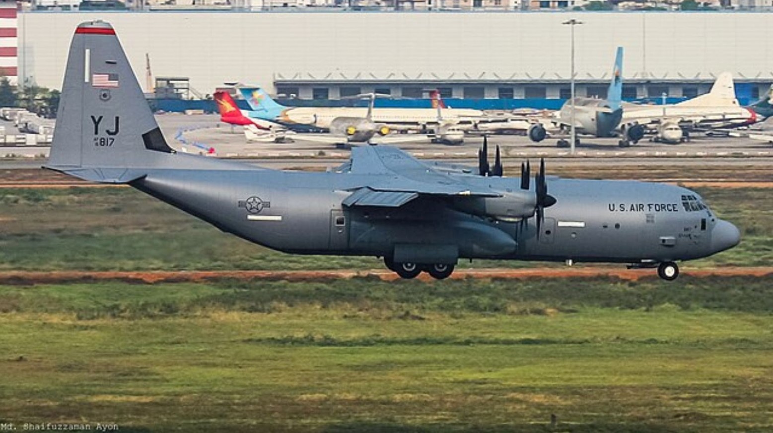 An image of a US Air Force plane landing next to a runway. Behind the plane is an airport runway with planes on.