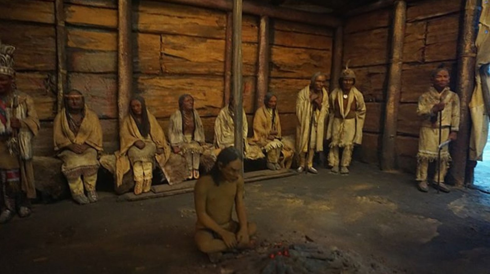 A recreation of an Iroquois tribe in a museum. One person is sitting on the floor next to a fire, and the others are either sitting or standing against a wooden wall.
