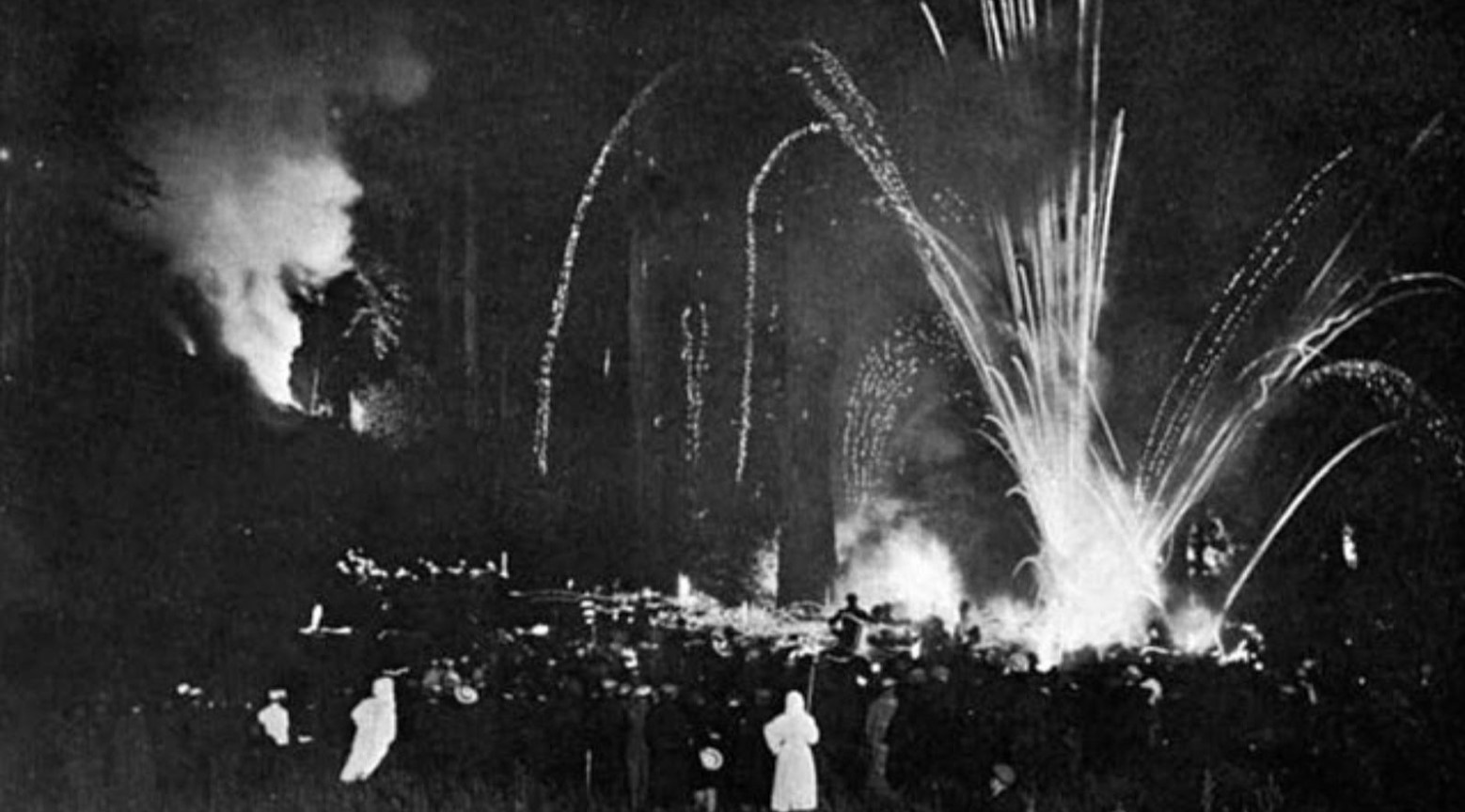 This is a black-and-white photo from the 1907 Cremation of the Care ceremony at Bohemian Grove. There is a crowd of people, and fireworks are going off.