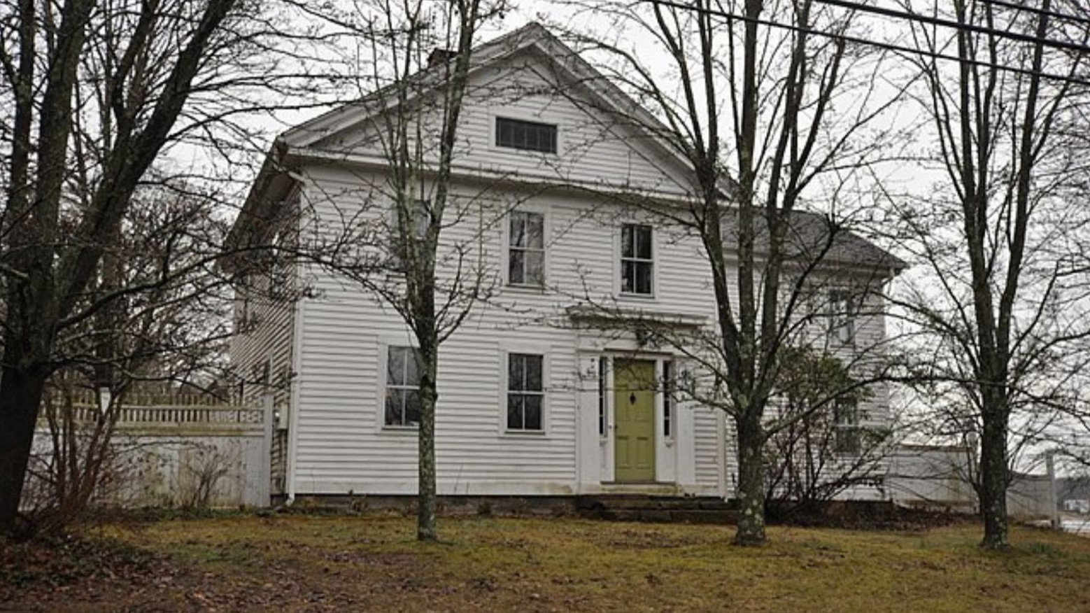 This is an old white house in Dudleytown, Connecticut. It has white pannelling and a green door and is surrounded by bare trees.