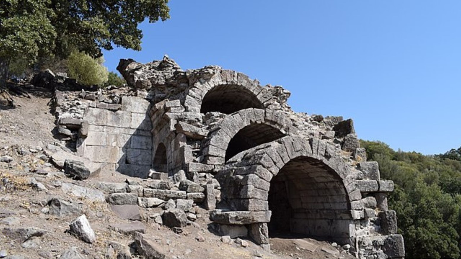 This is an ancient part of Aegae. Three stone tunnels are on top of one another on a hill. Green trees and bushes are on either side of the tunnels.