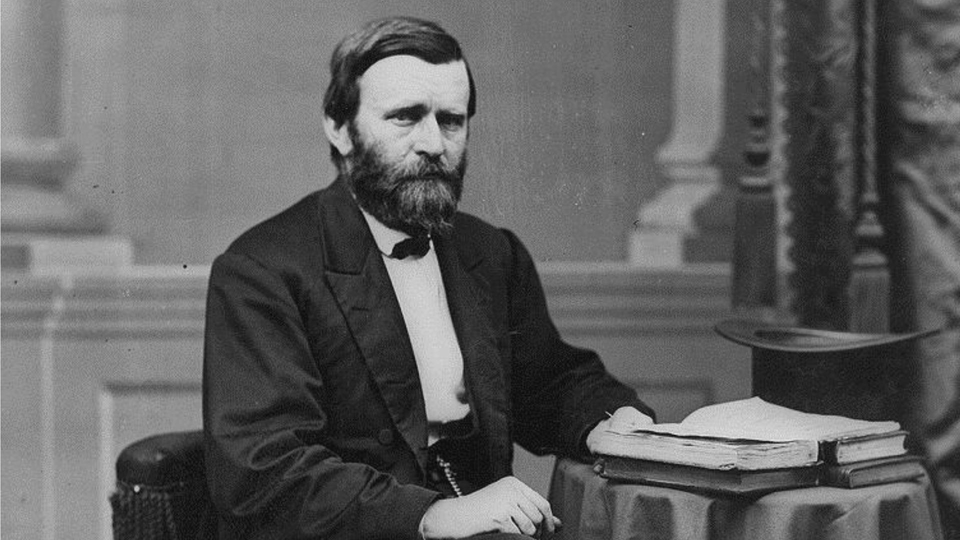A black and white photograph of Ulysses S. Grant, the 18th President of the United States, seated next to a table with documents. He is dressed in a dark suit with a bow tie