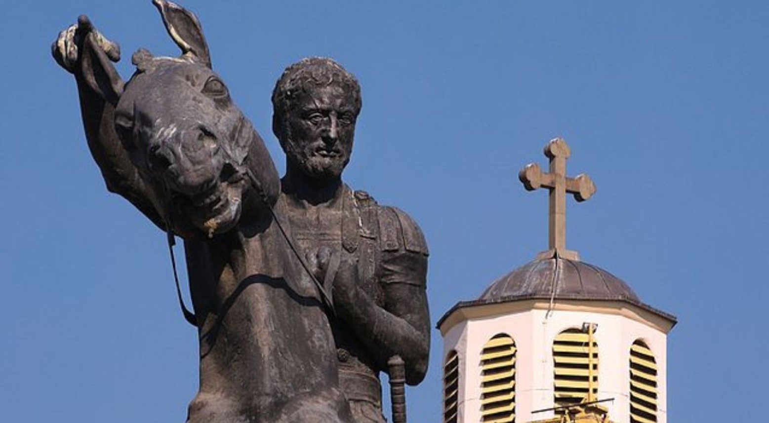 A statue of Philip II on a horse. There is a white tower in the background with a cross on top.