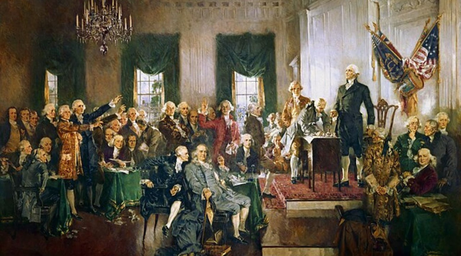 A painting of the signing of the US Constitution. A crowd is sitting and standing in the room, with one man standing up on a platform at the front. The US flag is on the wall behind him.