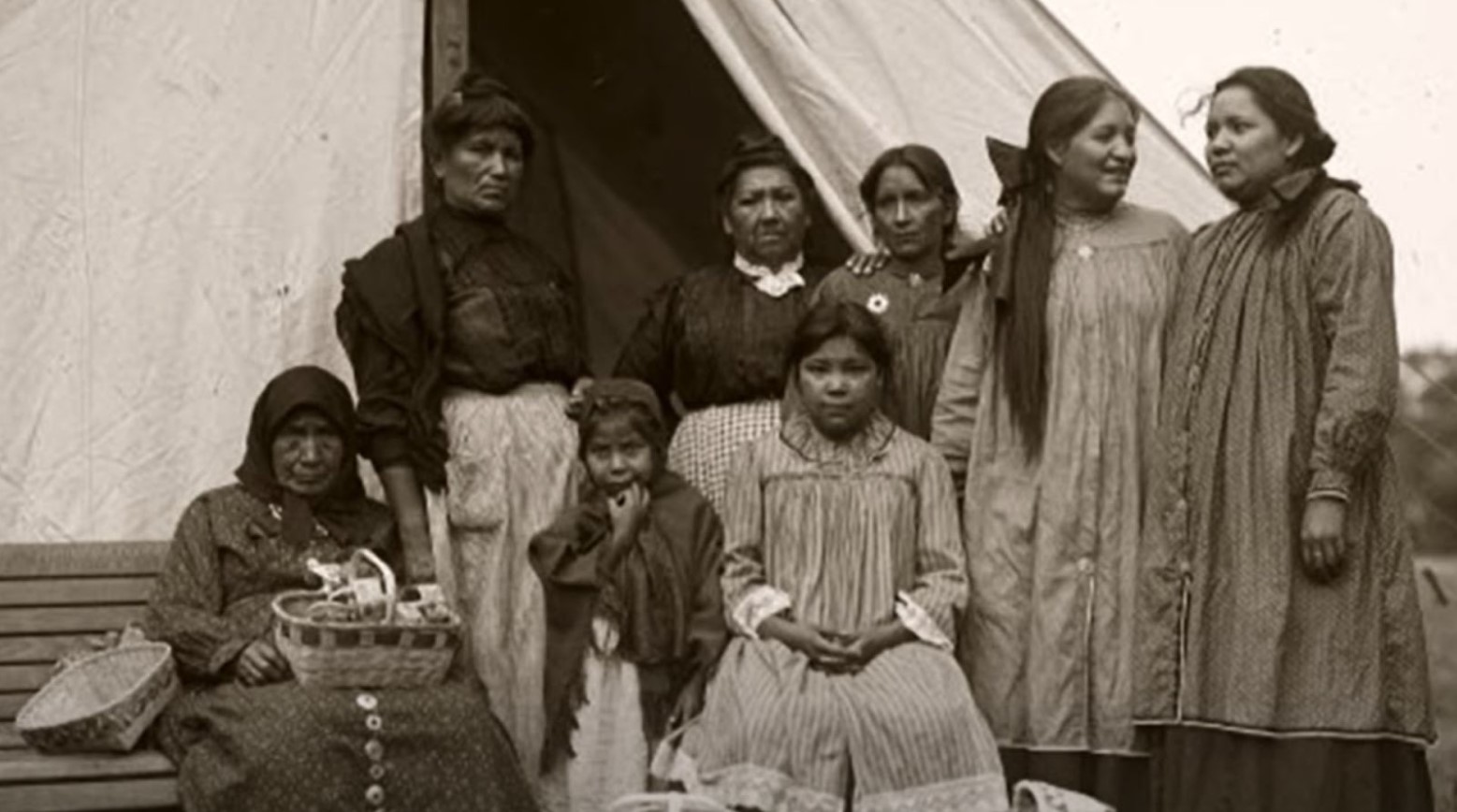 This is a black-and-white photo of some Iroquois women. They are all either standing or sitting in front of a tent.