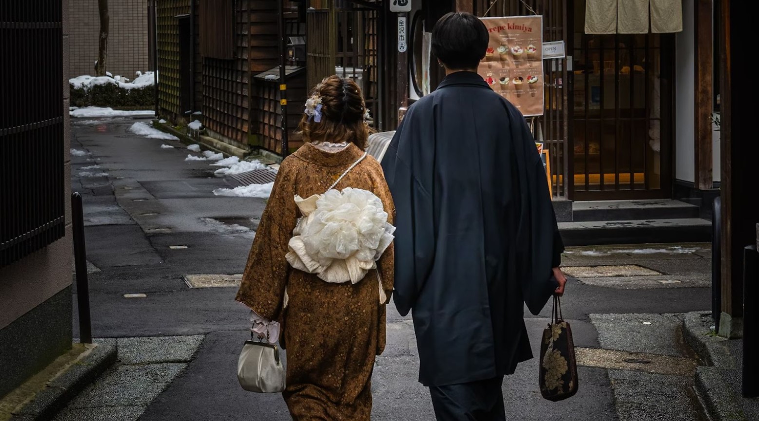 A man and woman walking down a street in the geisha district. The woman is wearing a yellow kimono and the man is wearing a grey coat. The street has some snow on it.