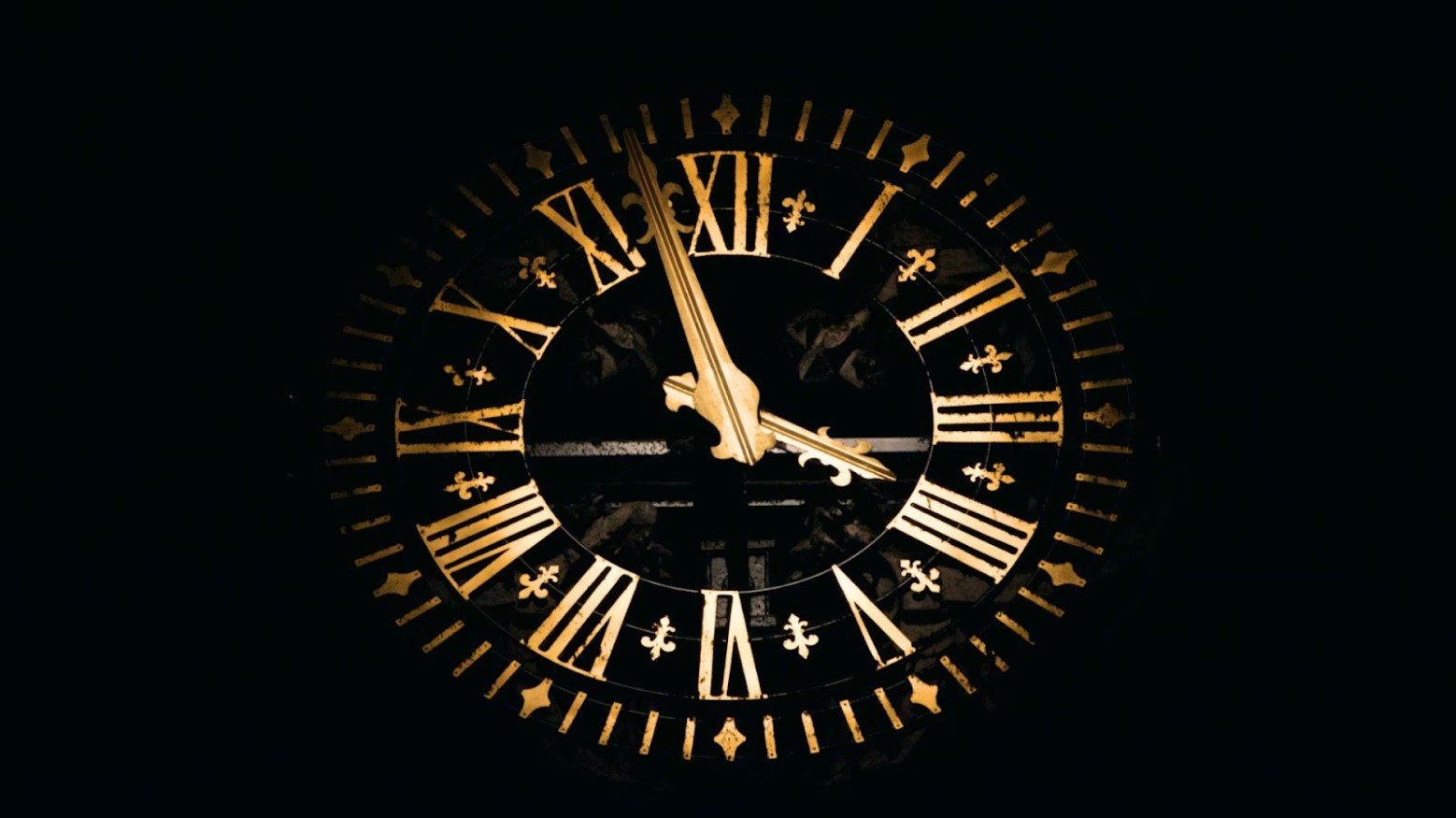 An old-fashioned clock with golden hands and numbers against a black background. The numbers are in Roman numerals from 1 to 12. The hands are pointing to 3:55.
