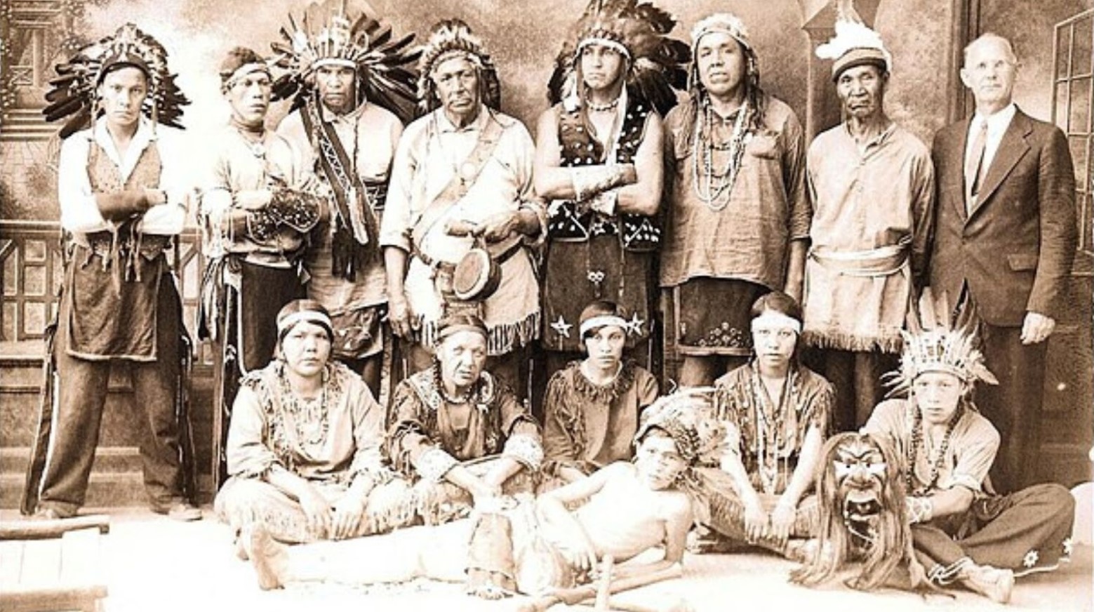 A black and white photo of a Native American tribe. They are all wearing traditional Native American dress, with one white man standing next to them in a suit.