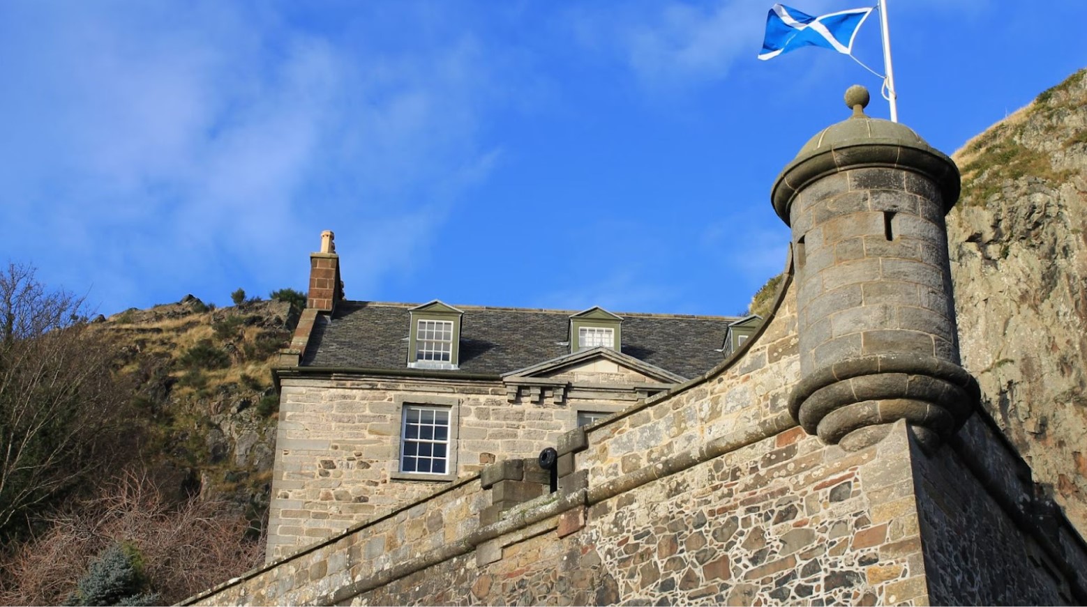 This is a photo of an old brick and stone building. At the top of the building is a white flagpole with the Scottish flag on it.