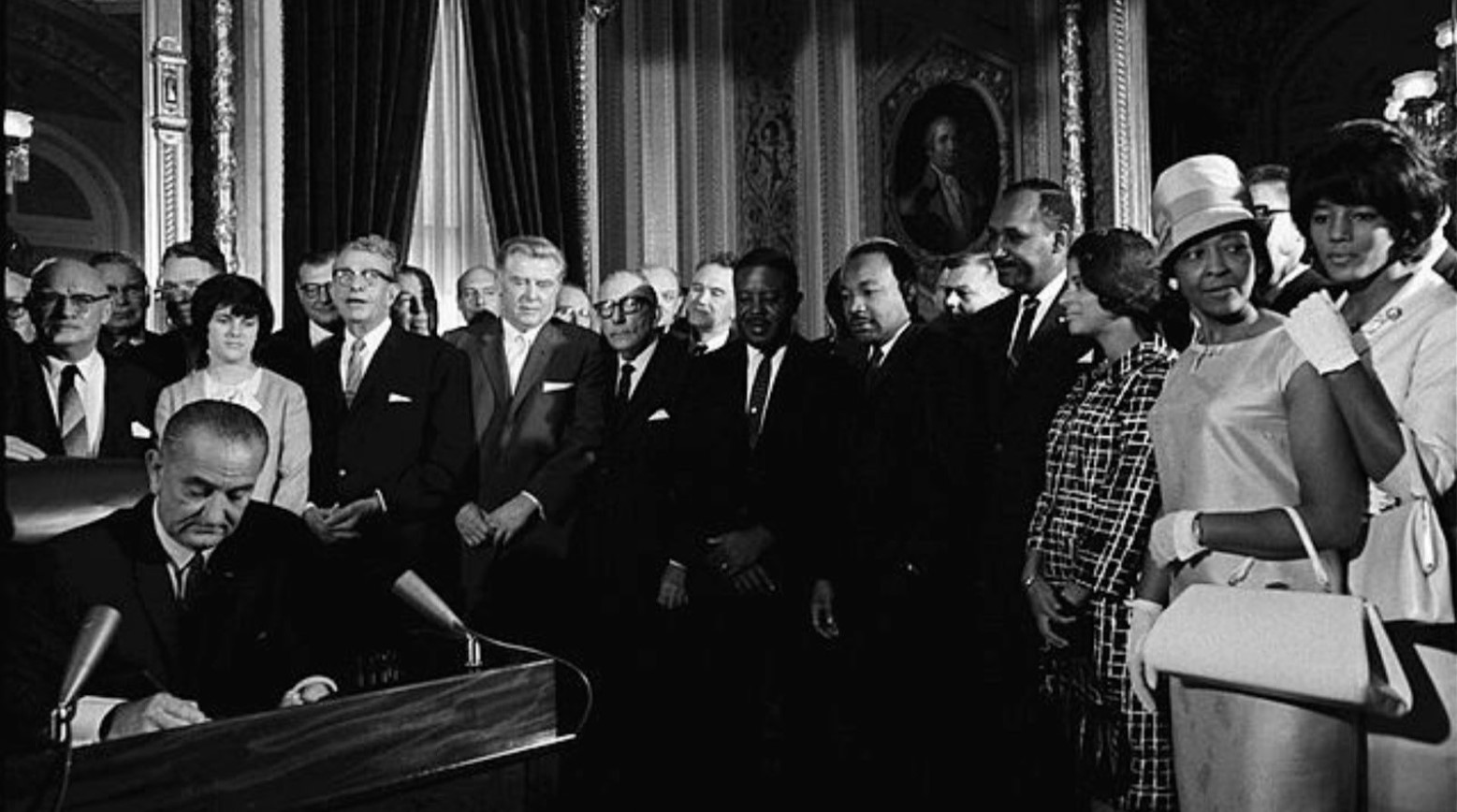 This is a black-and-white photo of the 1965 Voting Rights Act signing. The image shows President Lyndon Johnson sitting at a desk signing the document, with a crowd of men and women standing around him.