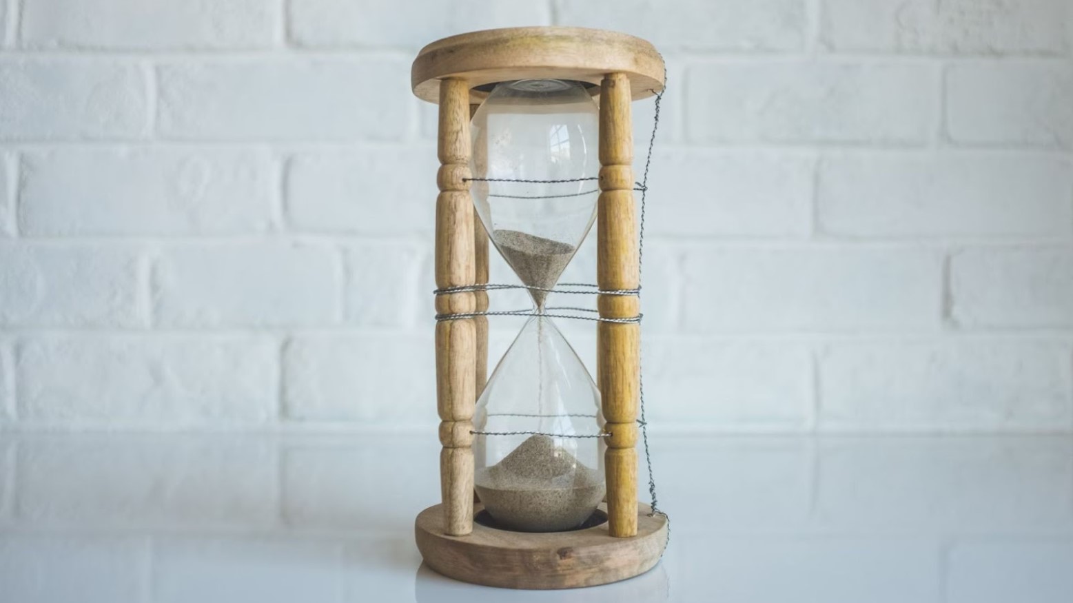An hourglass timer with sand in it. The hourglass has a wooden frame and clear glass. There is sand in the hourglass, with there being more sand at the bottom than at the top.