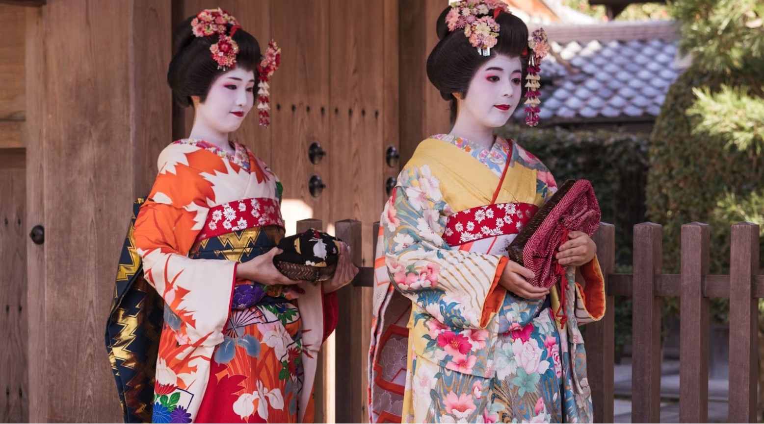 A pair of geishas. They are both in traditional geisha kimonos, with brightly colored floral kimonos and white-painted faces.